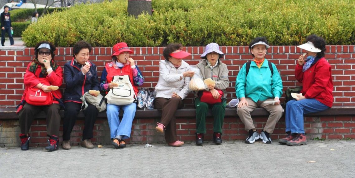 middle-aged asian women sitting in a row on the public bench, eating and sharing food. They wear colourful mismatched clothes with backpacks and hats.