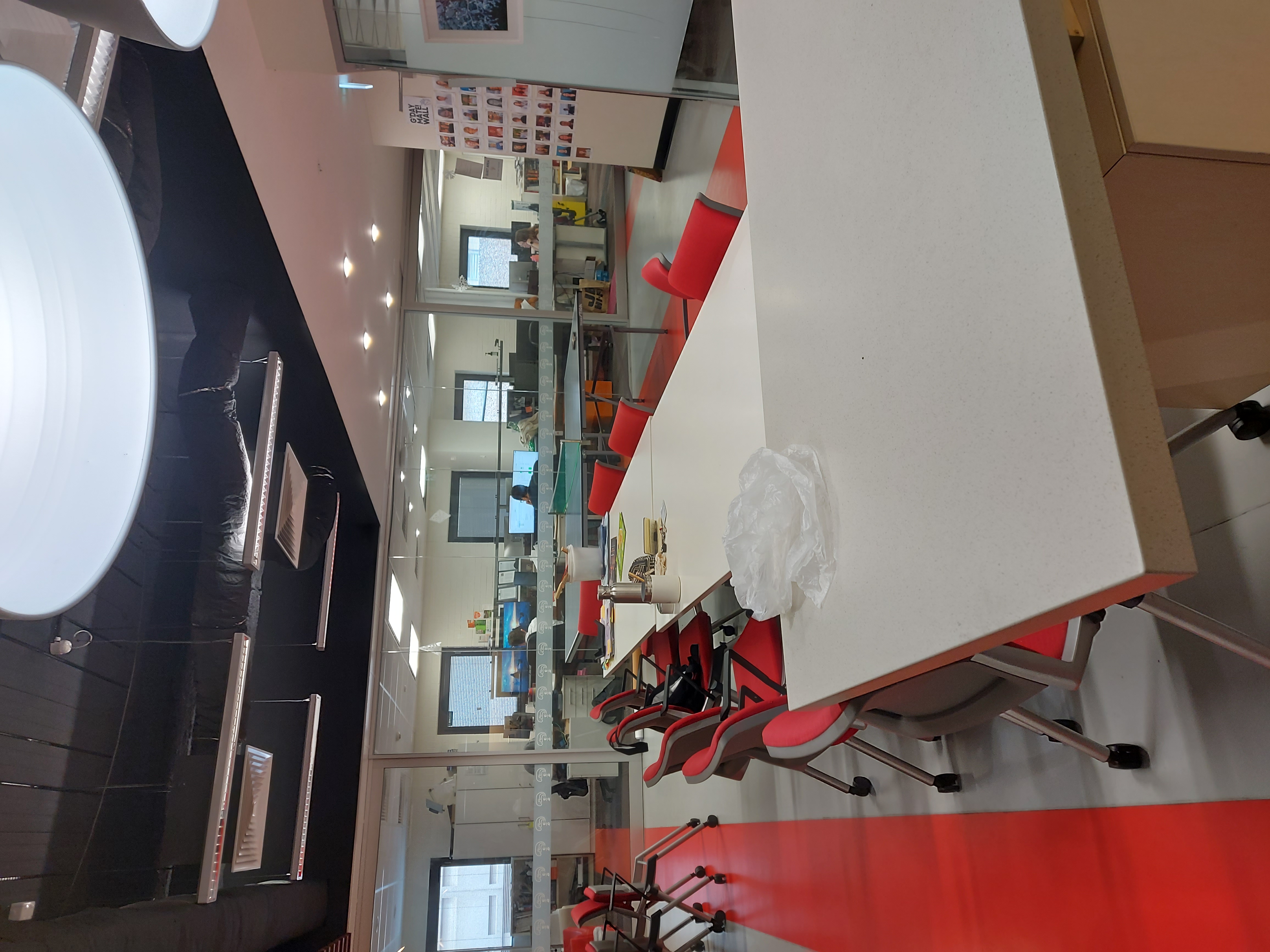 A large long white table with red chairs on a cozy grey and red patterned carpeted floor. In the background, there are rows of computers.