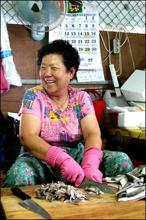 Ajumma smiling whilst cleaning fish. She wears colourful tight short sleeved t-shirt and loose fitting clothes, squatting on the floor.